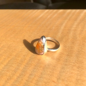 May include: A teardrop-shaped opal ring with a silver band. The opal is a mix of orange, pink, and white.