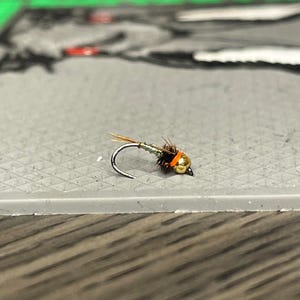 May include: A small, brown and gold fly fishing lure with an orange bead. The lure is tied on a hook and is ready to be used for fishing.