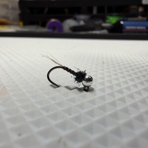 May include: A fishing fly lure featuring a curved metal hook with a dark brown segmented body, a small cluster of black synthetic fibers near the eye, and a shiny silver metallic bead head attached to the front of the hook shank.