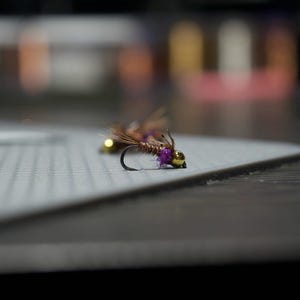 May include: Two brown and gold fly fishing lures with purple accents on a gray surface.