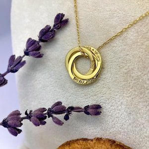 Peut inclure: Un collier doré avec trois anneaux entrelacés, chacun gravé d'un nom, du mot « love » et d'une date. Le collier est suspendu à une fine chaîne. Des brins de lavande pourpre et une tranche d'orange séchée sont en arrière-plan.