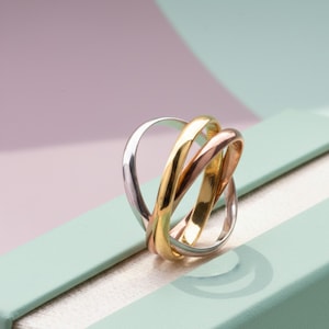 Peut inclure: Une bague à trois anneaux entrelacés, en argent, or et or rose. Posée sur une surface bleu clair, avec un fond rose et vert doux, elle offre un contraste attrayant. Ce bijou est un accessoire élégant et intemporel.
