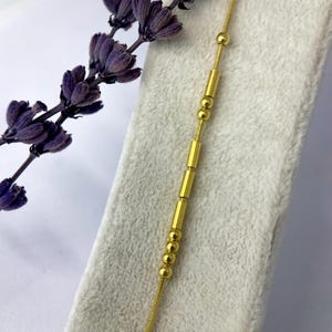 以下が含まれることがあります： 小さな丸いビーズと長方形のバーが連なった繊細なゴールドカラーのネックレス。 ネックレスは、乾燥した紫色のラベンダーの小枝の隣の白い表面に表示されています。 チェーンは細く、全体的なデザインはミニマリストです。