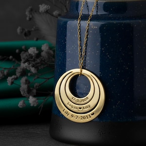 May include: A gold pendant necklace featuring three concentric circular rings. Each ring is engraved with a name and date. The names are Logan, Amy, and Lily. The dates are 12.3.2008 and 9-7-2011. The necklace is suspended from a fine chain.