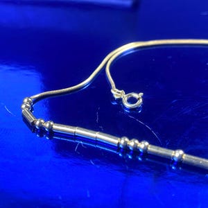 May include: A gold-toned necklace with a delicate chain and a series of small, round beads interspersed with longer, cylindrical beads. The necklace has a simple clasp and is set against a vibrant blue background.