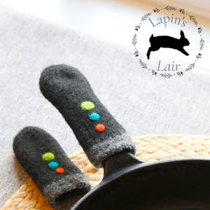 Peut inclure: Manchons de poignée de casserole en tricot gris foncé avec des pois colorés. Les manchons sont conçus pour s'adapter aux poignées d'une poêle en fonte. L'image comprend également un logo avec le texte "Lapin's Lair" et une silhouette de lapin.