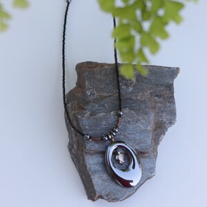 May include: A black beaded necklace with a large, oval-shaped pendant featuring a small turtle design. The pendant is made of a dark, metallic material and has a shiny finish. The necklace is displayed on a grey rock.