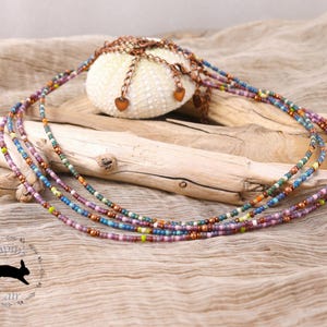 May include: Several multi-strand beaded necklaces in purple, blue, green, and copper tones are displayed over driftwood and a white sea urchin. The necklaces have small, colourful beads and heart-shaped charms. A logo with a rabbit silhouette is visible.