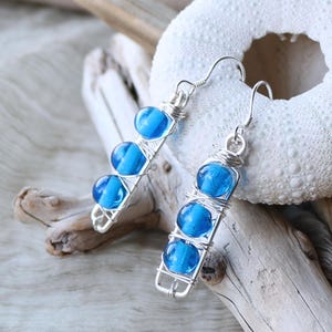Peut inclure: Une paire de boucles d'oreilles en fil d'argent avec des perles de verre bleues. Les boucles d'oreilles sont longues et pendantes, avec les perles disposées en ligne verticale.