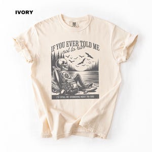 May include: A beige t-shirt with a black and white graphic of a skeleton sitting by a lake. The graphic includes the text "If you ever told me not to run, I'd still be standing next to you."