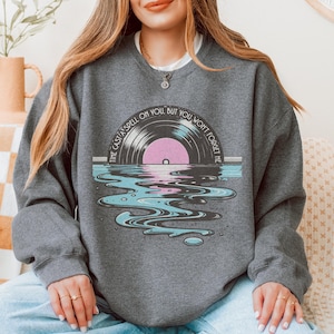May include: A dark gray sweatshirt featuring a graphic of a vinyl record with a pink center, set against a water reflection in shades of blue and gray. The text "TIME CAST A SPELL ON YOU, BUT YOU WON'T FORGET ME" is printed around the record.