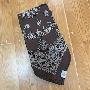 May include: A folded brown bandana with a white paisley pattern. The bandana has a small tag with a bee logo. The fabric is a dark brown color with a detailed white design.