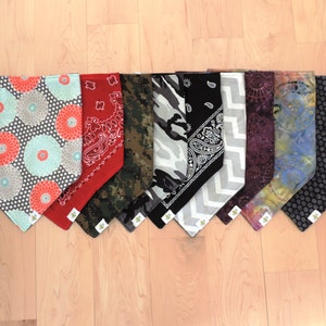 May include: A collection of nine dog bandanas in various patterns and colors. Designs include floral, paisley, camouflage, and polka dots. The bandanas are arranged on a light wood surface.