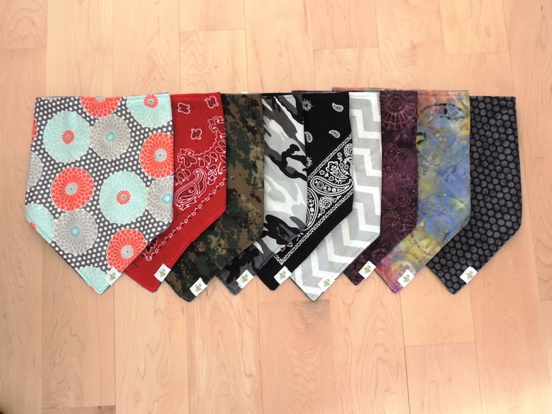 May include: A collection of nine dog bandanas in various patterns and colors. Designs include floral, paisley, camouflage, and polka dots. The bandanas are arranged on a light wood surface.