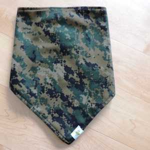 May include: A camouflage bandana in shades of green, brown, and black, with a pixelated digital pattern. The bandana has a triangular shape and a small white tag with a logo on the bottom corner.
