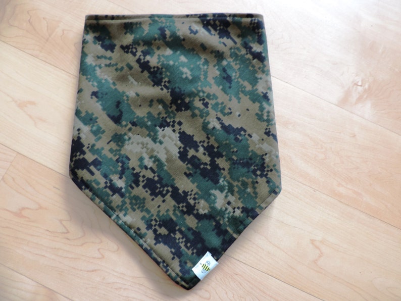 May include: A camouflage bandana in shades of green, brown, and black, with a pixelated digital pattern. The bandana has a triangular shape and a small white tag with a logo on the bottom corner.