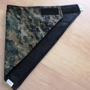 May include: A reversible pet bandana featuring a digital camouflage pattern in shades of green, brown, and tan. The reverse side is solid black. The bandana has a hook and loop closure for easy adjustment and a small tag with text.