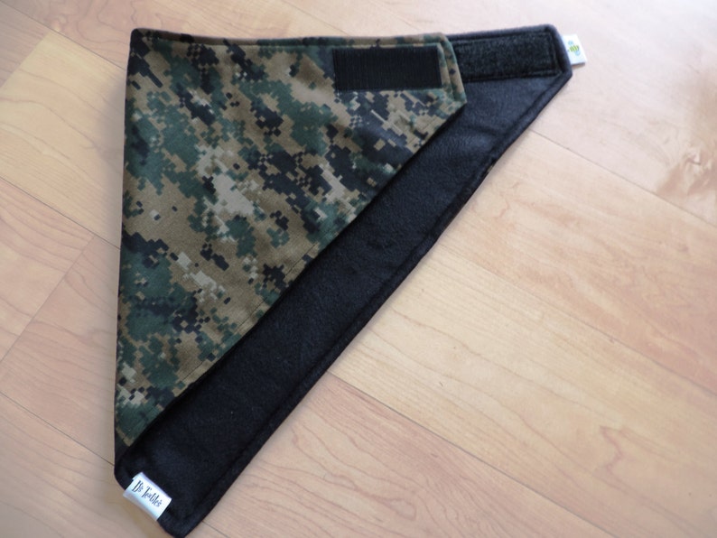 May include: A reversible pet bandana featuring a digital camouflage pattern in shades of green, brown, and tan. The reverse side is solid black. The bandana has a hook and loop closure for easy adjustment and a small tag with text.