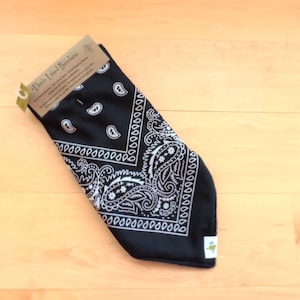 May include: A black fleece-lined bandana with a white paisley pattern. The bandana is folded and has a tag attached. The tag reads "Fleece Lined Bandana". The bandana is on a light wood surface.