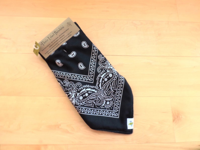 May include: A black fleece-lined bandana with a white paisley pattern. The bandana is folded and has a tag attached. The tag reads "Fleece Lined Bandana". The bandana is on a light wood surface.