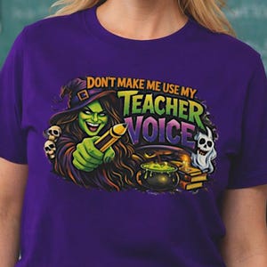 May include: A purple t-shirt featuring a cartoon witch with the text "DON'T MAKE ME USE MY TEACHER VOICE". The witch is holding a pencil and is surrounded by skulls and a cauldron. The background includes a chalkboard and a world map.