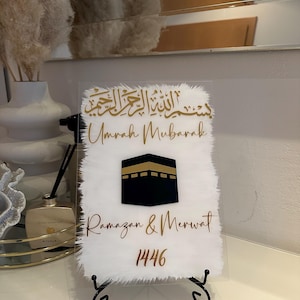 Könnte beinhalten: Ein weißes Acrylschild mit goldenen Buchstaben und einem schwarzen und goldenen Bild der Kaaba. Das Schild trägt die Aufschrift "Masjid Al Haram", "Umrah Mubarak", "Ramazan & Merwat" und "1446".