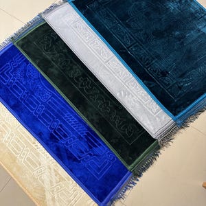 Könnte beinhalten: Fünf Gebetsteppiche mit Fransen an den Rändern. Die Teppiche sind in verschiedenen Farben: beige, blau, grün, weiß und türkis. Jeder Teppich hat ein kompliziertes Design mit arabischer Kalligraphie.