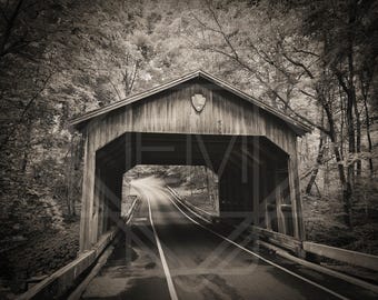 Könnte beinhalten: Sepiafarbene Fotografie einer überdachten Brücke mit einer Straße, die hindurchführt. Die Holzbrücke ist von Bäumen umgeben, wodurch ein Tunneleffekt entsteht. Ein Wappen ist auf dem Dach der Brücke zu sehen.