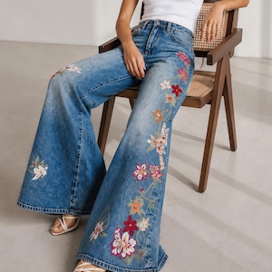 Op de afbeelding: Een wit geribd hemdje gecombineerd met een blauwe denim broek met wijde pijpen en kleurrijke bloemenborduursels op de benen. De jeans heeft een hoge taille en een wijd uitlopend silhouet, versierd met rode, roze, oranje en witte bloemmotieven.