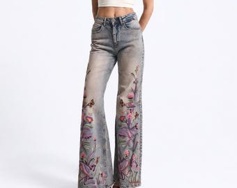 May include: A cream-colored short-sleeved crop top paired with light-wash blue denim jeans featuring a wide-leg cut. The jeans are decorated with colorful floral and butterfly embroidery patterns extending from the hem up to the mid-thigh area.