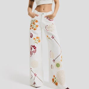 Może przedstawiać: Białe dżinsy z szerokimi nogawkami z kolorowym haftem kwiatowym. Haft przedstawia pomarańczowe, czerwone i zielone kwiaty oraz inne elementy dekoracyjne. Modelka ma na sobie biały top.