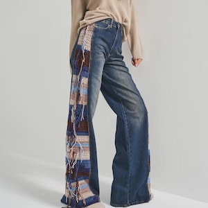 Könnte beinhalten: Blaue Jeans mit weitem Bein und einem einzigartigen Häkeldesign auf den Seiten. Die Häkelpaneele weisen eine Mischung aus Braun-, Blau- und Beigetönen mit Fransen auf. Das Model trägt einen beigen Pullover und eine braune Mütze.