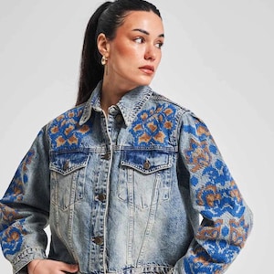 May include: A light blue denim jacket with embroidered floral details in blue and gold thread. The jacket has a classic collar, button closure, and two front pockets.
