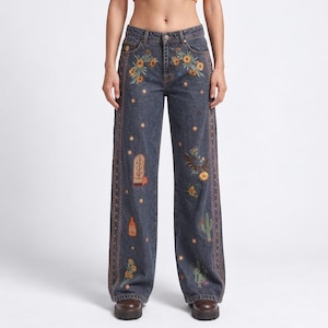 Op de afbeelding: Een oranje geribbelde tanktop gecombineerd met een donkergrijze spijkerbroek met geborduurde bloemen-, cactus- en hemelse motieven. De broek heeft een rechte pasvorm met decoratieve zijstiksels en een knoopsluiting.