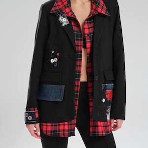 May include: Black blazer with red and black plaid lining and collar. The blazer features denim pocket patches and decorative embellishments. The outfit includes a red and black plaid shirt and black trousers. The blazer has a unique, artistic design.
