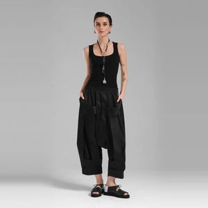 May include: A black sleeveless tank top paired with black wide-leg trousers featuring prominent vertical fabric straps and a low-slung crotch. The outfit is accessorized with a long black beaded necklace and black open-toe sandals.