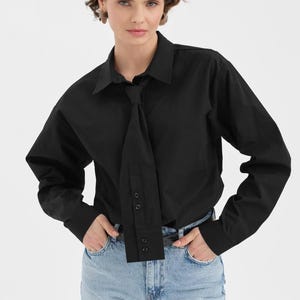 Puede incluir: Una camisa negra de manga larga con un dise&ntilde;o &uacute;nico tipo corbata. La camisa presenta un cuello cl&aacute;sico y pu&ntilde;os abotonados. La modelo lleva vaqueros azul claro. La camisa est&aacute; hecha de una tela lisa y de color s&oacute;lido.
