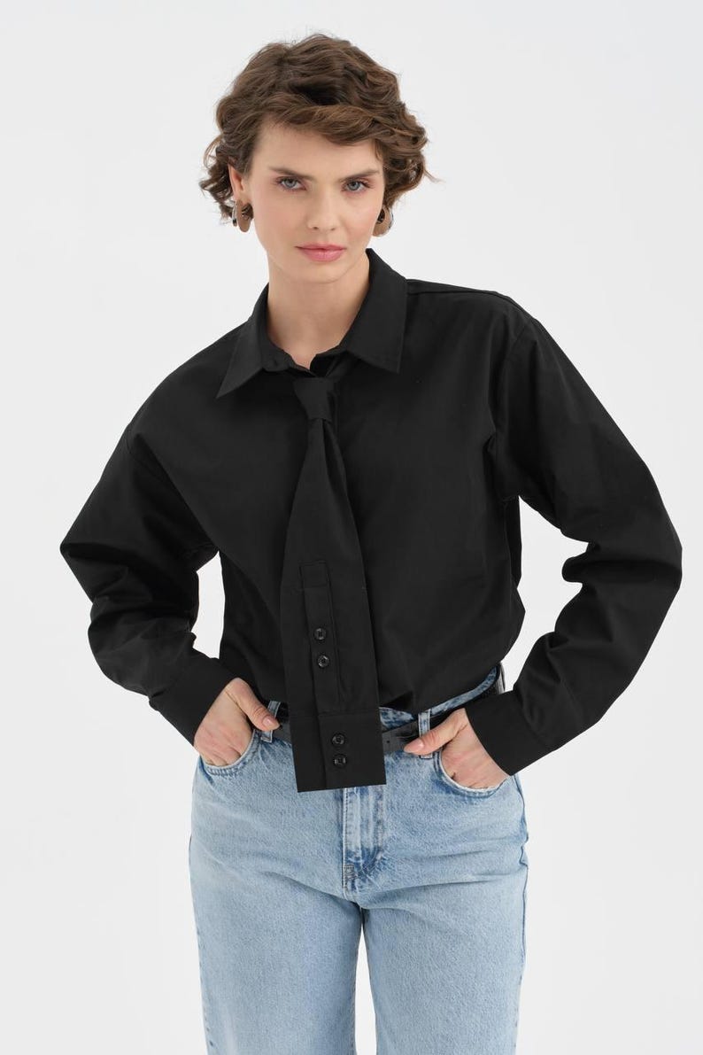 Puede incluir: Una camisa negra de manga larga con un dise&ntilde;o &uacute;nico tipo corbata. La camisa presenta un cuello cl&aacute;sico y pu&ntilde;os abotonados. La modelo lleva vaqueros azul claro. La camisa est&aacute; hecha de una tela lisa y de color s&oacute;lido.