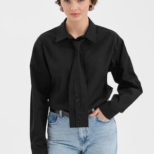 Puede incluir: Una camisa negra con un dise&ntilde;o &uacute;nico tipo corbata. La camisa de manga larga tiene cuello y botones. La modelo lleva vaqueros azul claro y un cintur&oacute;n negro. El elemento tipo corbata a&ntilde;ade un toque moderno a un estilo cl&aacute;sico.