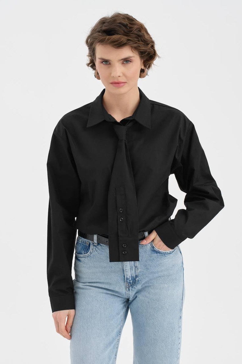 Puede incluir: Una camisa negra con un dise&ntilde;o &uacute;nico tipo corbata. La camisa de manga larga tiene cuello y botones. La modelo lleva vaqueros azul claro y un cintur&oacute;n negro. El elemento tipo corbata a&ntilde;ade un toque moderno a un estilo cl&aacute;sico.