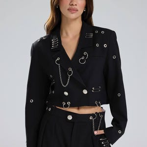 May include: Black cropped blazer adorned with silver safety pins, chain details, and ring accents. The double-breasted blazer features silver buttons and is paired with matching black trousers. A contemporary, edgy fashion piece.