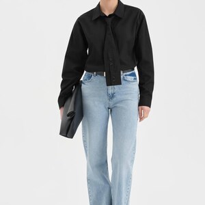 Puede incluir: Una camisa negra de manga larga con detalle de corbata, combinada con vaqueros azul claro. El conjunto se completa con gafas de sol negras y un clutch negro. Los vaqueros tienen un dise&ntilde;o de pernera ancha.
