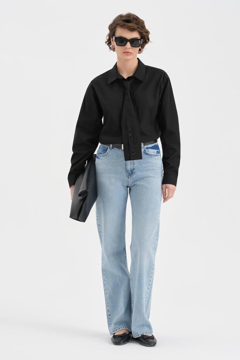 Puede incluir: Una camisa negra de manga larga con detalle de corbata, combinada con vaqueros azul claro. El conjunto se completa con gafas de sol negras y un clutch negro. Los vaqueros tienen un dise&ntilde;o de pernera ancha.