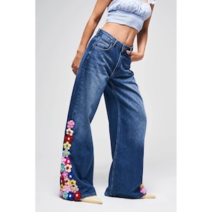 Könnte beinhalten: Blaue Jeans mit weitem Bein und farbenfrohen floralen Häkeldetails an der Seite der Beine. Die Jeans haben ein klassisches Fünf-Taschen-Design und einen Knopf- und Reißverschluss. Die Jeans werden mit einem hellblauen Crop-Top getragen.