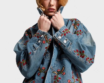 May include: A light blue denim jacket featuring red and green floral embroidery. The jacket has a classic design with button fastenings and pockets. The embroidery is detailed and adds a decorative element to the garment.