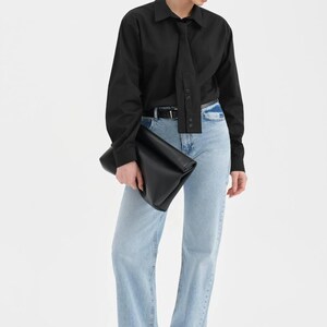 Puede incluir: Una persona con una camisa negra con detalle de corbata, vaqueros azul claro y bailarinas negras. Tambi&eacute;n lleva gafas de sol negras y sostiene un clutch negro. El atuendo es sencillo y elegante.