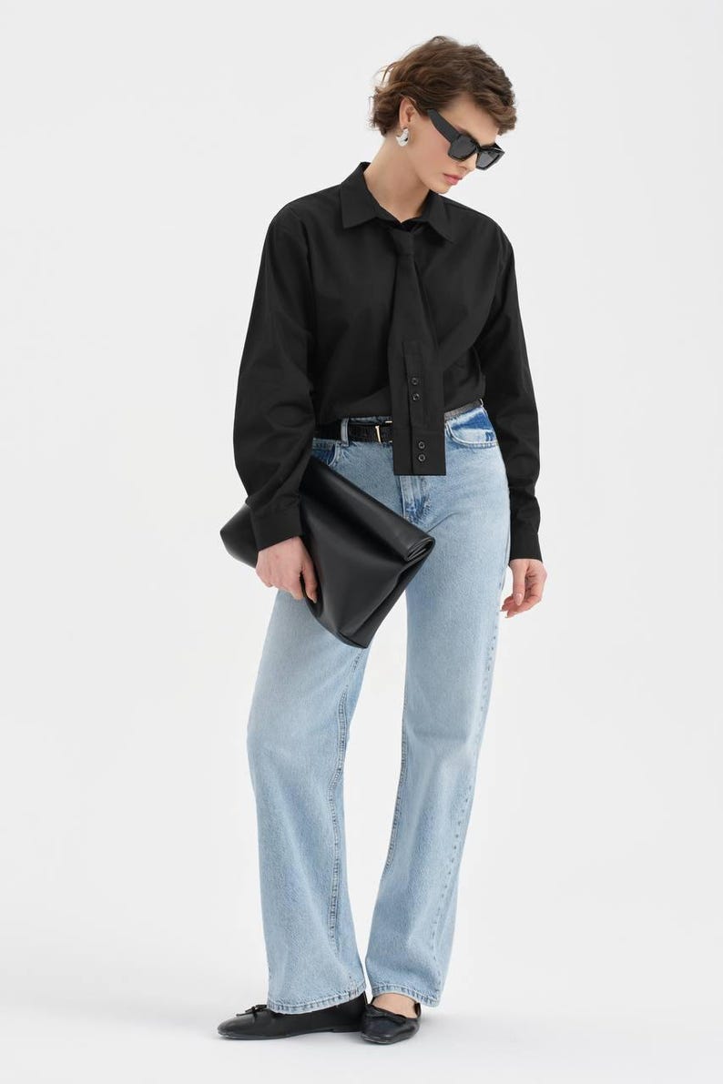Puede incluir: Una persona con una camisa negra con detalle de corbata, vaqueros azul claro y bailarinas negras. Tambi&eacute;n lleva gafas de sol negras y sostiene un clutch negro. El atuendo es sencillo y elegante.