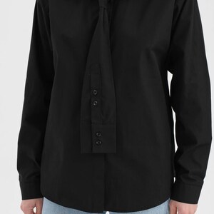 Puede incluir: Una camisa negra de manga larga con un dise&ntilde;o &uacute;nico tipo corbata. La camisa tiene un cuello cl&aacute;sico y una serie de botones en el detalle tipo corbata. La camisa se combina con vaqueros azul claro. Una prenda elegante y moderna.