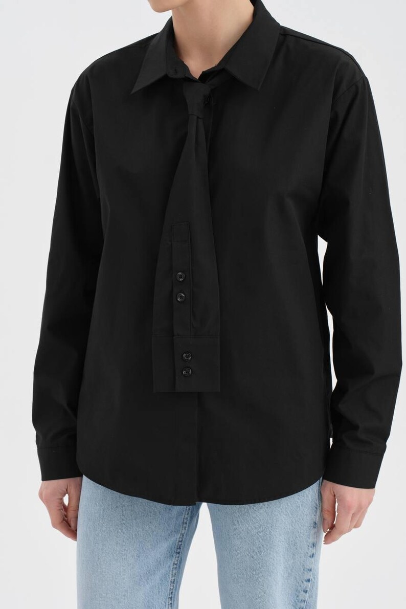 Puede incluir: Una camisa negra de manga larga con un dise&ntilde;o &uacute;nico tipo corbata. La camisa tiene un cuello cl&aacute;sico y una serie de botones en el detalle tipo corbata. La camisa se combina con vaqueros azul claro. Una prenda elegante y moderna.