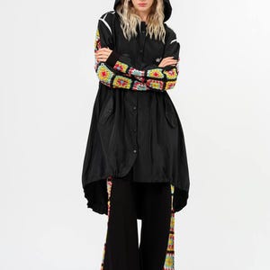 May include: Black hooded jacket with white trim and colorful crochet sleeves. The jacket is paired with black pants featuring crochet detailing down the sides. The outfit is a blend of modern and bohemian styles.